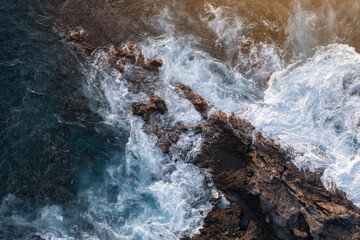 Aerial view of ocean waves crashing breaking against coastline rocks. Seascape ocean background from above. Blue water splashing and foaming at cliffs in sunset time. Landscape of Tenerife, Canary