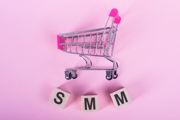 SMM - Social media marketing, word on wooden cubes, on a pink background with a shopping trolley.