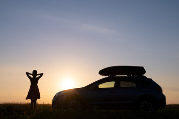 Dark silhouette of woman driver standing near her car on grassy field enjoying view of bright sunset. Young female relaxing during road trip beside SUV vehicle