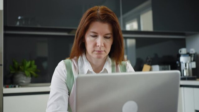 Female employee with long hair wearing shirt and vest working on laptop from home, typing and looking at screen, glossy furniture and kitchen devices. Concept of remote work and outsource