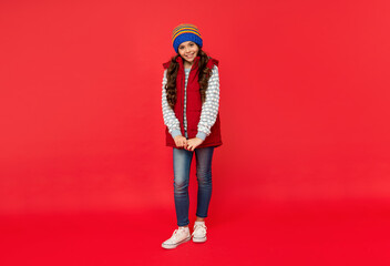 full length of happy teen girl in knitted winter hat and down vest on red background, winter outfit