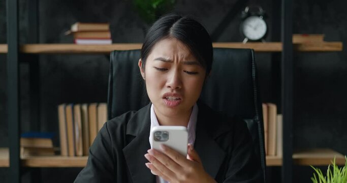 Sad upset pensive asian businesswoman looking at smartphone, say no, unpleasantly surprised and scared while sitting at office desk. Shocked young female looking at mobile phone reaction on lose