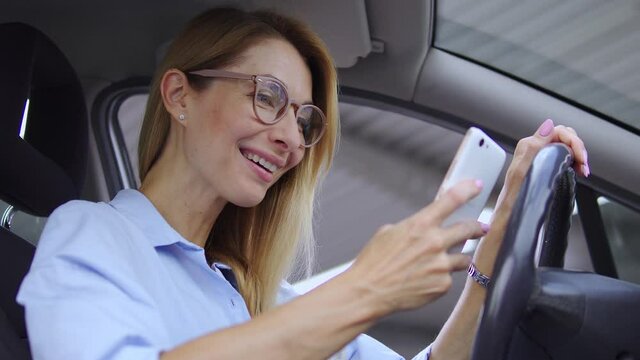 Excited blond woman in modern eyeglasses sitting in her car, receiving message on smartphone, surprised with great sale and discounts offer, has many coupons for good shopping, black Friday