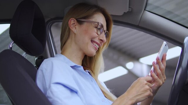 Attractive pleased woman in stylish eyeglasses with blond hair smiling sitting on driver seat of her car and reading good news on smartphone, satisfied with information about free car maintenance