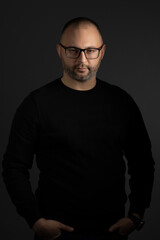 A young man in black-rimmed glasses and a black sweater keeps his hands in his pockets posing for...