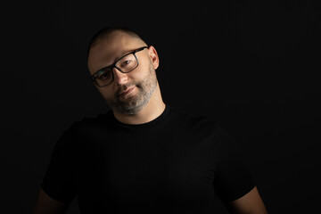 Fototapeta premium A young man in a black T-shirt with a short haircut and black-rimmed glasses poses against a dark background carefully and aggressively looking at the camera