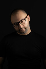 Obraz premium A young man in a black T-shirt with a short haircut and black-rimmed glasses poses against a dark background carefully and aggressively looking at the camera