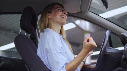 Joyful blond lady in casual shirt dancing active and positive dance sitting on driver seat in her car, enjoying music and moving quickly, celebrating own business start-up opening, success