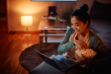 Happy Asian woman eats popcorn while watching movie on laptop at night at home. - obrazy, fototapety, plakaty