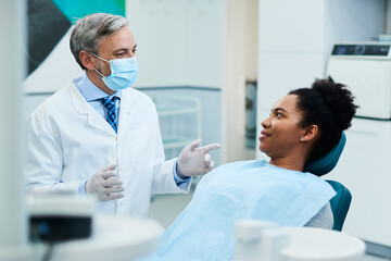 Obraz premium Dentist with face mask talks to black female patient during appointment at dentist's office.