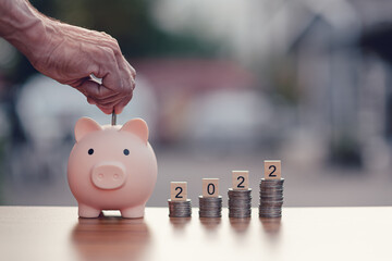 Hand putting coin in to pink piggy bank and coins stack and Text of 2022 number written on wooden...
