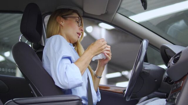 Blond woman in casual shirt and stylish eyeglasses sneezing closing mouth with tissue sitting in car, catching cold or flu, suffering seasonal allergy, allergy reaction on odor or dust