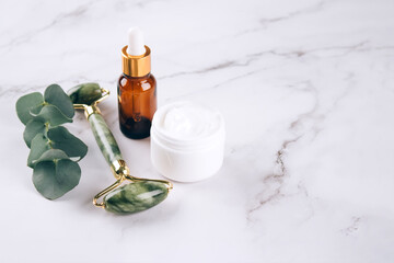 Jade facial gua sha roller with beauty serum, face oil and face cream on white marble table background with copy space. Facial massage kit for lifting massage therapy made from natural stones. Mock up