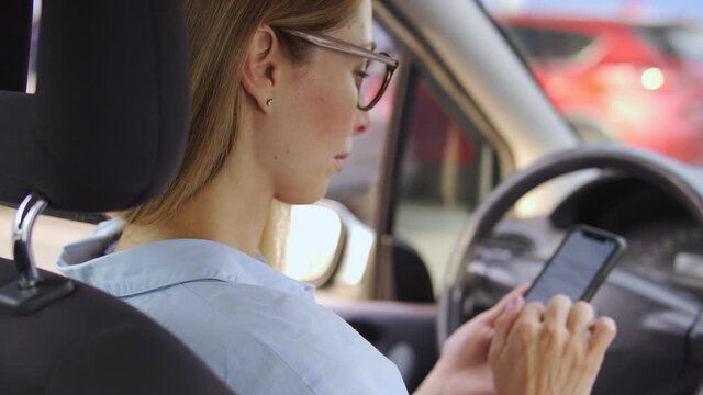 Concentrated business woman in elegant eyeglasses looking through documents online on smartphone app sitting on driver seat in car, preparing to important meeting with partners or clients