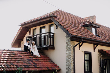 Guy in a suit, white shirt and black hat, and a girl in a wedding dress and a white hat, are sitting on the roof of the house holding hands. Young couple on the wedding day on the roof of the house