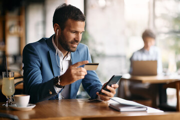 Smiling businessman uses cell phone and credit card for online shopping while enjoying in cafe,