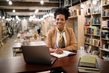 Happy black university teacher holds online class from library and looks at camera. - obrazy, fototapety, plakaty