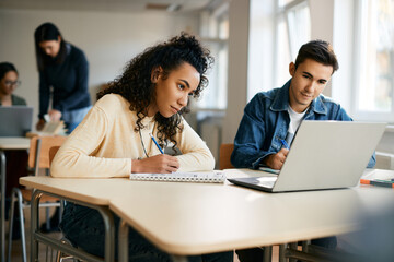 High school classmates take notes while e-learning on laptop in classroom.