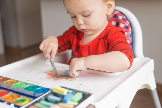 18 Month Old Toddler Drawing And Painting With Two Hands At Once; Watercolor Paint And Chalk Pastel Being Used While Seated In High Chair