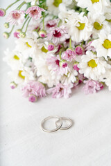 Fototapeta na wymiar Pair of wedding rings on a white surface with beautiful white and purple flowers in the background. Commitment and love concept. Vertical orientation