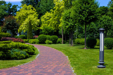 lantern iron ground garden lighting of park curved path paved with stone tiles in backyard among plants, evergreen bushes and foliage trees surrounded by green lawn on sunny summer day, nobody.