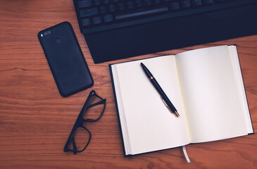 Workplace. On a wooden table are located: keyboard, phone, glasses, notebook and pen.