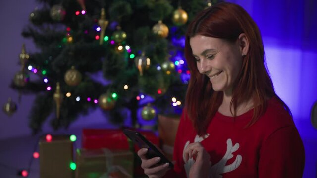 happy girl with mobile phone on new year's eve, young woman shows at the camera a smartphone with a green screen and a gift on the background of a Christmas tree with many lights at night