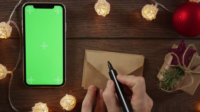 congratulations with winter holidays concept: present gift craft box and smartphone with green screen on wooden desk. Christmas new year background lighting garland, red ornament ball and cell phone