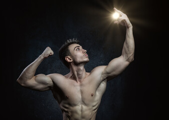 Obraz premium Young male athlete bodybuilder aestheticist posing against a dark background with sunlight in his hand. Healthy lifestyle and sports concept.