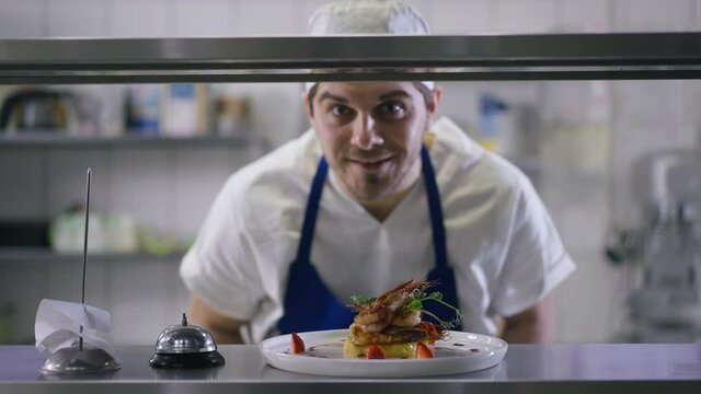Rack focus from confident smiling cook putting plate with grilled prawn ringing service bell to delicious fried seafood on countertop. Positive Caucasian chef serving tasty dinner in restaurant posing