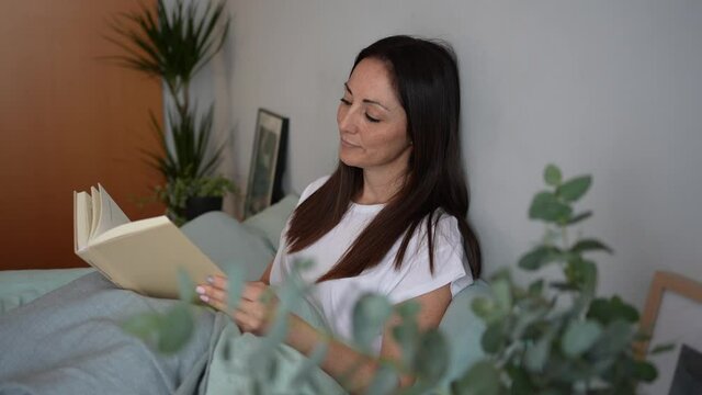  A beautiful woman reading a book and relaxing on the bed in the bedroom in the morning. Resting on the weekend with a book in a calm environment.