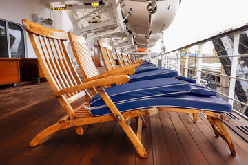 Deckchairs, with blue foldec cushions, lined up on a cruise line on a cloudy port day. Liferafts can be seen hanging above the deck. With space for text. - 477528712
