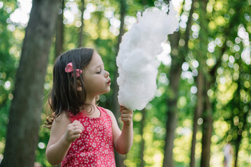 Little cute girl 3-4 eating cotton candy in sunny park among tall trees on green grass
