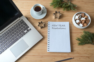 New Year Resolutions written on a spiral notebook on a desktop with laptop, coffee, cookies and fir branches, copy space, top view from above