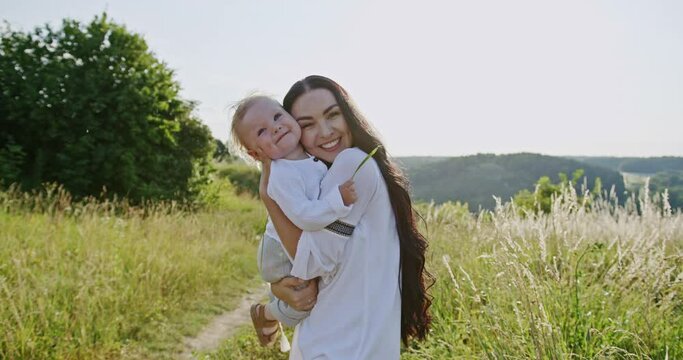 Beautiful family portrait of a loving mother with her baby both smiling happily at the camera outdoors in a rural landscape as she hugs the infant to her cheek