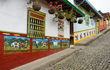 Fototapeta na wymiar Guatape, Colombia: Calle del Recuerdos is a cobblestone pedestrian street that passes between colorful tile-roofed buildings decorated with zocalos (friezes) and hanging pots of flowers