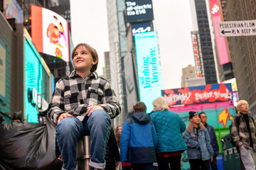 boy in jeans and plaid jacket sitting on post in new york city, time square