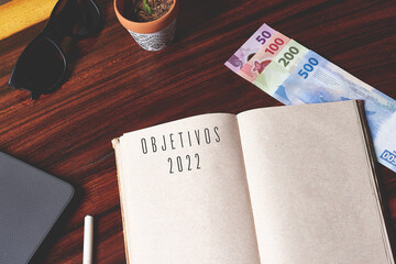 Blank notebook with Mexican money, with glasses on a wooden desk and a gray laptop with a...