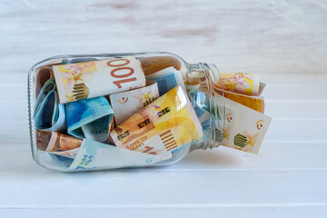 Israeli shekels in glass jar over white wooden background.