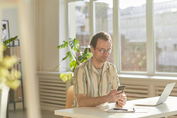 Horizontal medium shot of handsome Caucasian man wearing eyeglasses sitting at desk in modern office surfing Internet on smartphone