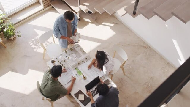 Top-view shot of four Asian architects working in team while discussing housing project looking at its paper layout sitting around table in bright modern office