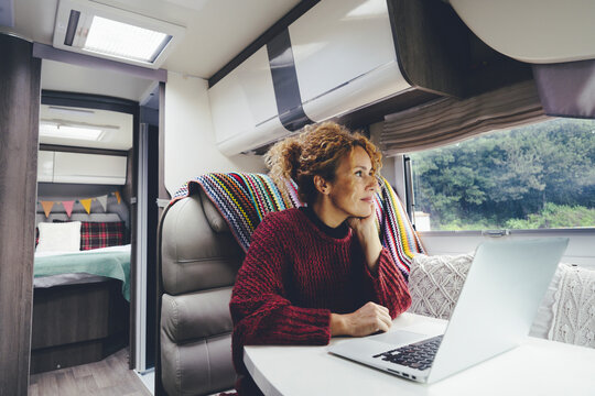 Adult woman use laptop computer inside a camper van recreational vehicle sitting at the table with bedroom in background and nature park outside the windos. Concept of travel and remote worker people