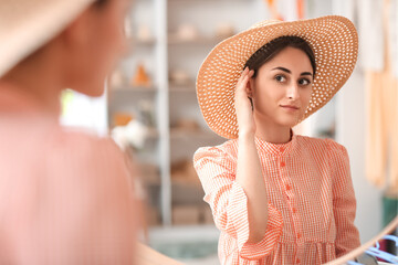 Young woman trying on straw hat near mirror in boutique