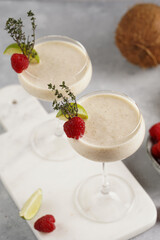 Cocktail glasses with sweet banana milk shake beverage, raspberries and coconut on white marble board