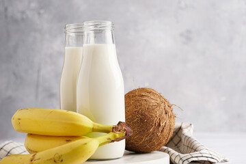 Two bottles with non-dairy milk, coconut and bananas on round marble board, grey background