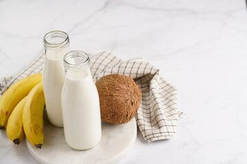 Two bottles with non-dairy milk, coconut and bananas on round marble board, grey background