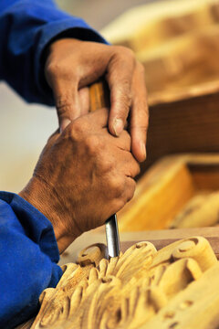A vertical close up detail shot of African male woodworker carving an intricate floral pattern with a chisel, into a piece of wood in a furniture factory, Johannesburg, South Africa