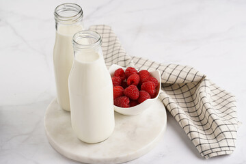 Two bottles with non-dairy milk and raspberries on round marble board, grey background