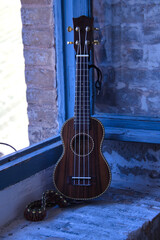 Brown colored ukulele with strap leaning against a brick window frame. Concept instruments, music,...