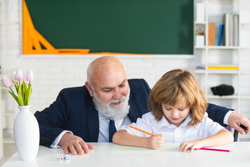 Senior teacher or grandfather and school boy pupil in classroom at school. Elementary pupil reading and writing with teacher in classroom.